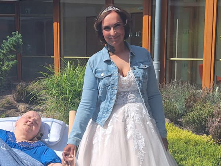 Braut Christina im Hochzeitskleid hält im sonnigen Innenhof des Albertinen Hospiz Norderstedt die Hand ihrer im Bett liegenden Mutter.