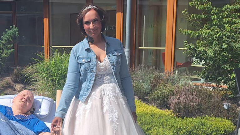 Braut Christina im Hochzeitskleid hält im sonnigen Innenhof des Albertinen Hospiz Norderstedt die Hand ihrer im Bett liegenden Mutter.