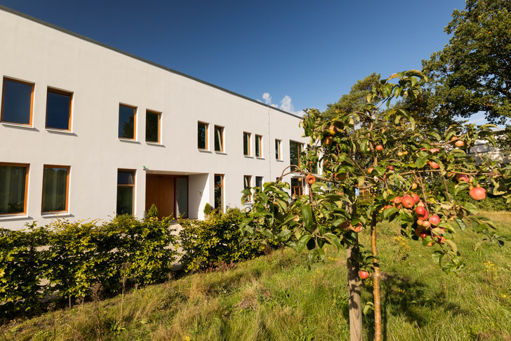 Seitliche Gebäudeansicht des Albertinen Hospiz Norderstedt mit einem Apfelbaum mit Äpfeln, Gartenhecke und Grünstreifen unter wolkenfreiem Himmel
