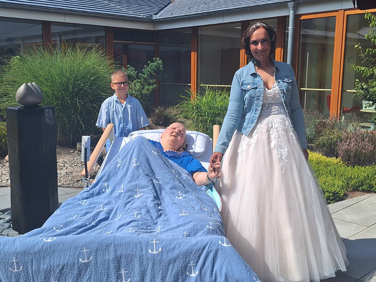 Gästin des Albertinen Hospiz Norderstedt hält im sonnigen Innenhof die Hand ihrer Tochter im Hochzeitskleid; ihr Enkel steht hinter dem Bett.