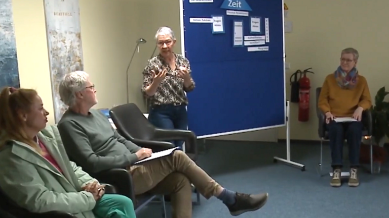 Kursleiterin Sabine Bornhöft steht an einer Pinnwand und spricht mit den im Kreis sitzenden Teilnehmenden beim Kurs für den ehrenamtlichen mobilen Hospizdienst im Albertinen Hospiz Norderstedt. Kursleiterin Sabine Bornhöft steht an einer Pinnwand und spricht mit den im Kreis sitzenden Teilnehmenden beim Kurs für den ehrenamtlichen mobilen Hospizdienst im Albertinen Hospiz Norderstedt.
